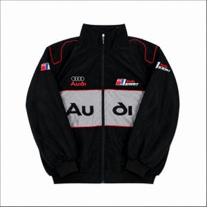 Może przedstawiać: Czarna kurtka zapinana na zamek z wysokim kołnierzem i szerokim szarym poziomym pasem na piersi. Kurtka posiada logo Audi oraz napis Audi Sport wyhaftowany w kolorach czerwonym, białym i czarnym z przodu oraz na rękawach z czerwoną lamówką.