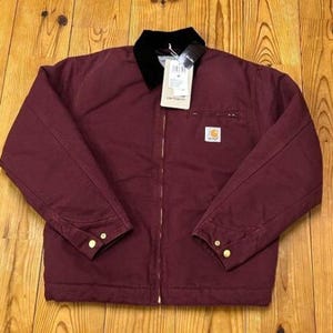 Könnte beinhalten: Burgunderfarbene Arbeitsjacke mit schwarzem Kragen und Reißverschluss. Die Jacke hat eine kleine Brusttasche und goldfarbene Knöpfe an den Manschetten. Ein weißes Etikett ist am Kragen befestigt.