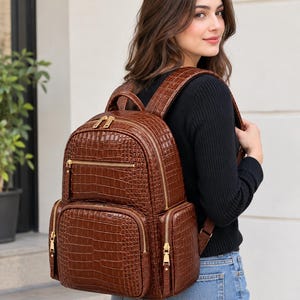 Può includere: Uno zaino marrone con texture a rilievo effetto coccodrillo, dotato di molteplici scomparti con cerniera e finiture color oro, una maniglia superiore e spallacci regolabili, progettato per trasportare oggetti personali.