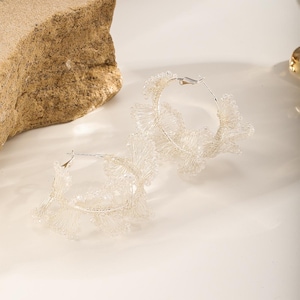 May include: A pair of silver hoop earrings with a delicate, floral-like design. The earrings feature a series of small, clear beads and wire, creating a textured, airy appearance. The earrings are displayed on a white surface.