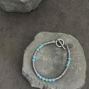 Puede incluir: Pulsera plateada con cuentas de color turquesa y cierre de palanca. La pulsera está hecha de metal enrollado y presenta pequeñas cuentas rectangulares de color turquesa espaciadas uniformemente alrededor de la banda. El texto en la parte superior dice: I DON'T WANT THIS FEELING TO BE REPRODUCED IN MY MEMORY.