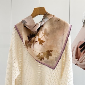 May include: A square scarf with a floral design in shades of beige, brown, and purple. The scarf is draped over a cream-coloured jumper. The scarf has a purple border and is hanging on a wooden hanger.