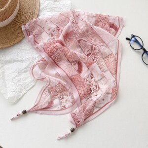May include: A light pink square scarf with a patchwork design featuring floral and geometric patterns. The scarf has a delicate, sheer appearance and is adorned with small wooden beads at the corners. A straw hat and glasses are also visible.