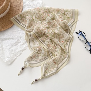 May include: A light green and cream-colored silk scarf with a floral pattern and a striped border. The scarf has decorative wooden beads at the ends. A straw hat and eyeglasses are also in the image.