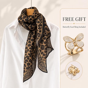 Puede incluir: Una bufanda con estampado de leopardo marrón y negro sobre una camisa blanca con cuello. La imagen incluye el texto FREE GIFT y Butterfly Scarf Ring Included, junto a un anillo para bufanda de metal dorado con forma de mariposa y alas esmaltadas.