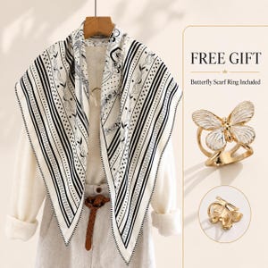 May include: A square scarf featuring a black and white pattern of stripes, stars, and floral motifs, draped over a cream-colored long-sleeve top. Included is a gold-toned butterfly scarf ring with white enamel details, labeled FREE GIFT.