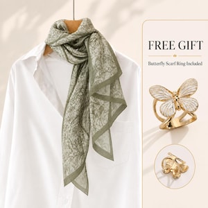 May include: A sage green scarf with a dark green floral pattern draped over a white collared shirt. To the right, a gold-toned metal scarf ring shaped like a butterfly with white enamel wings is displayed, along with a smaller view of the ring.