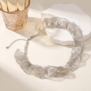May include: A silver-colored necklace with a ruffled design. The necklace has a delicate, layered appearance, with a chain and clasp closure. The necklace is displayed on a white surface, with a glass and a white decorative object in the background.