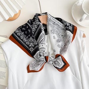 May include: A square scarf with a paisley pattern in black and white, trimmed with a brown border. The scarf is tied in a knot and displayed on a white top.