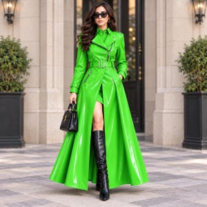 Puede incluir: Una gabardina de color verde lima vibrante hecha de un material brillante tipo charol. El abrigo presenta una solapa ancha, un cinturón con hebilla, herrajes plateados, bolsillos con cremallera y una falda larga y fluida. Botas negras altas.