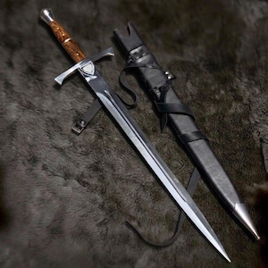 Può includere: Una spada in acciaio con lama lucida, una guardia con un dettaglio centrale a forma di scudo e un manico in legno venato con pomolo metallico. La spada è accompagnata da un fodero in pelle nera con finiture in metallo color argento.
