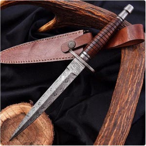 May include: A Damascus steel dagger with a patterned blade and a brown leather-wrapped handle. The dagger is accompanied by a brown leather sheath. The handle has a metal crossguard. The knife is displayed on a black surface with a wooden log and antler.