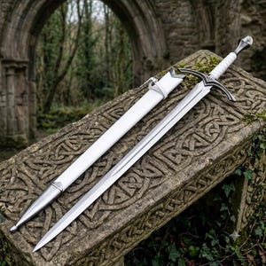 Op de afbeelding: Een zilverkleurig metalen zwaard met een puntig lemmet en een centrale groef. Het gevest heeft een wit omwikkelde greep, een pareerstang met decoratieve metalen accenten en een afgeronde pommel. Een bijpassende witte schede met zilveren metalen beslag.