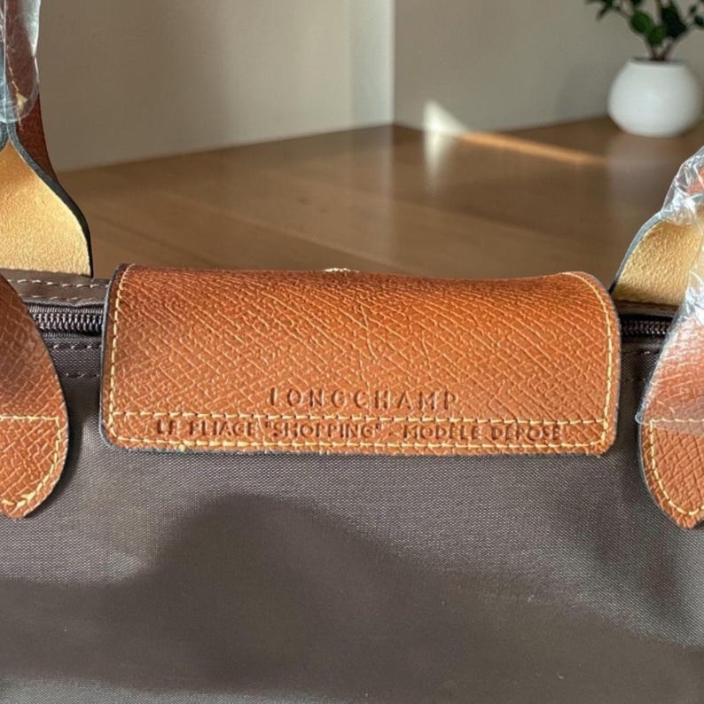 Op de afbeelding: Bruine handtas met een leren handvat en een bruine leren flap. Op de flap staat de merknaam "LONGCHAMP" en de tekst "LE PLIAGE SHOPPING MODELE DEPOSE".