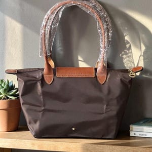 Op de afbeelding: Een bruine tote bag met leren handvatten en details. De tas heeft een rechthoekige vorm met een ritssluiting en een kleine flap met de merknaam. De handvatten zijn verpakt in doorzichtig plastic.