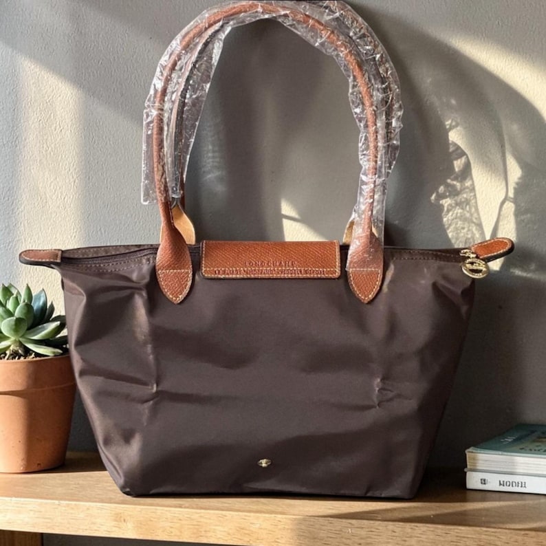 Op de afbeelding: Een bruine tote bag met leren handvatten en details. De tas heeft een rechthoekige vorm met een ritssluiting en een kleine flap met de merknaam. De handvatten zijn verpakt in doorzichtig plastic.