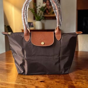 Op de afbeelding: Een bruine tote bag met een leren flap en handvatten. De tas heeft een goudkleurige knoop op de flap en een ritssluiting. De handvatten zijn verpakt in doorzichtig plastic.
