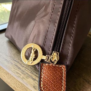 Op de afbeelding: Close-up van een bruine nylon handtas met een goudkleurige rits en een goudkleurig cirkelvormig embleem. Het embleem toont een silhouet van een persoon op een paard. Een bruin leren bandje is aan de rits bevestigd.