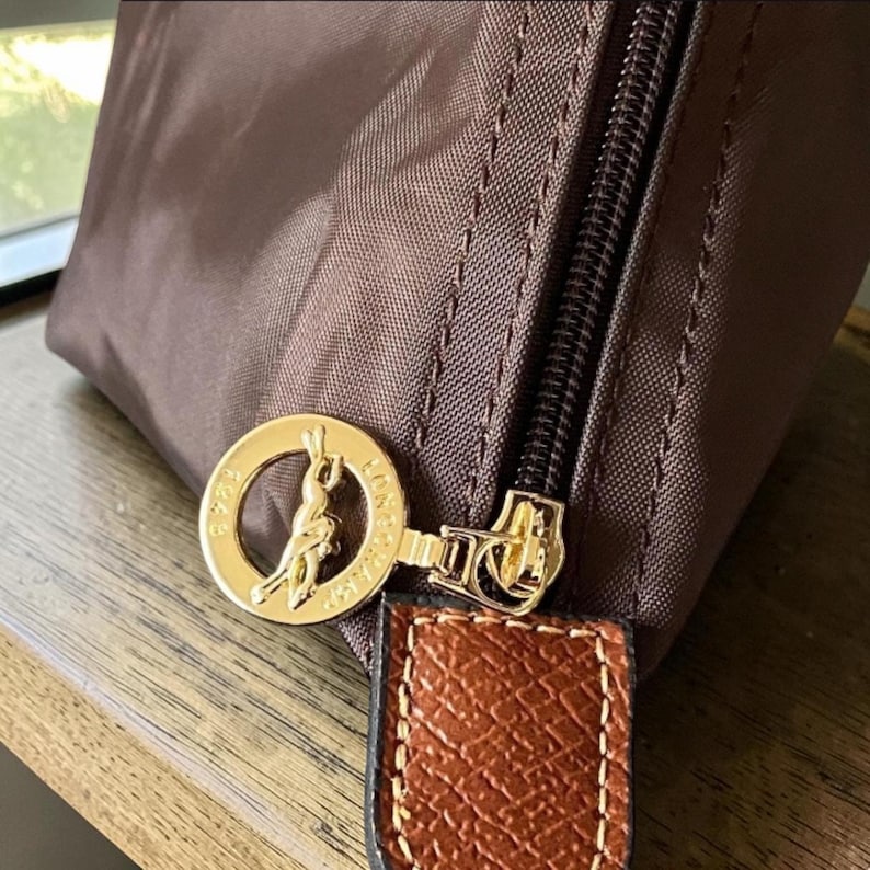 Op de afbeelding: Close-up van een bruine nylon handtas met een goudkleurige rits en een goudkleurig cirkelvormig embleem. Het embleem toont een silhouet van een persoon op een paard. Een bruin leren bandje is aan de rits bevestigd.