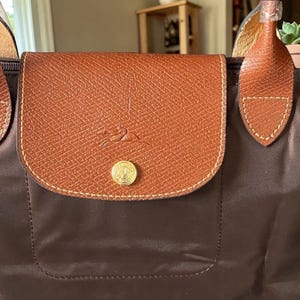Op de afbeelding: Een bruine tote bag met een klepsluiting. De klep is gemaakt van getextureerd bruin leer en heeft een goudkleurige knoop en een logo. De body van de tas is van een donkerdere bruine stof en de handvatten zijn van lichter bruin leer.