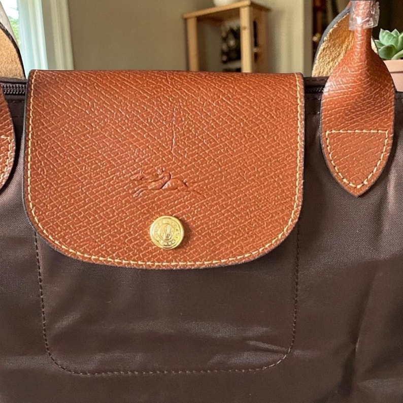 Op de afbeelding: Een bruine tote bag met een klepsluiting. De klep is gemaakt van getextureerd bruin leer en heeft een goudkleurige knoop en een logo. De body van de tas is van een donkerdere bruine stof en de handvatten zijn van lichter bruin leer.
