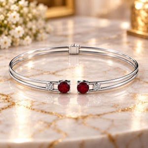Peut inclure: Un bracelet ouvert argenté doté de deux pierres précieuses rondes rouge foncé et de deux pierres plus petites, claires, rondes et facettées aux extrémités. Le bracelet présente un design à double anneau relié à l'arrière par une charnière.