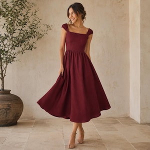 Puede incluir: Un vestido midi burdeos con escote cuadrado y mangas cortas. El vestido tiene un corpiño ajustado y una falda acampanada. La modelo lleva tacones color nude. El vestido está hecho de una tela suave y fluida.
