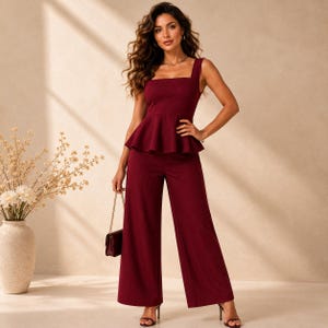 May include: A deep burgundy two-piece set consisting of a sleeveless peplum top with a square neckline and matching wide-leg trousers. The outfit features a solid, textured fabric and is paired with a small dark handbag with a gold chain strap.