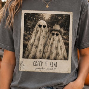 May include: A charcoal gray short-sleeve t-shirt featuring a rectangular graphic print of two ghosts wearing heart-shaped sunglasses and a cowboy hat. The print resembles a vintage polaroid with the text CREEP IT REAL and pumpkin patch.
