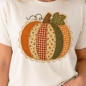 May include: A cream-coloured t-shirt featuring a large, central pumpkin graphic. The pumpkin is composed of various fabric-like patches in orange, green, and beige with floral, gingham, and polka dot patterns, bordered by a delicate cream lace trim.
