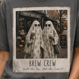 May include: A charcoal gray t-shirt featuring a rectangular graphic print of two figures dressed as ghosts wearing sunglasses and holding coffee cups in front of a cafe. The text reads "GHOST BEAN CAFE", "BREW CREW", and "Spill the Tea... Not the Cider".