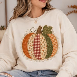 Peut inclure: Un sweat-shirt à col rond couleur crème arborant un grand motif central de citrouille. La citrouille est composée de divers empiècements en tissu aux motifs orange, vichy rouge et floraux verts, bordés d'une fine dentelle blanche.