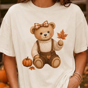 May include: A cream-colored short-sleeve t-shirt featuring a graphic of a brown teddy bear wearing a plaid bow, a beige sweater, and brown overalls. The bear holds an orange maple leaf, with a small pumpkin and fallen leaves at its feet.