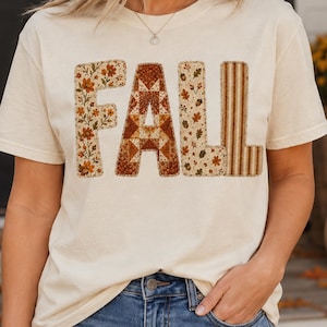 May include: A cream-coloured short-sleeve t-shirt featuring the word FALL printed across the chest. Each letter has a distinct pattern: the F has orange flowers, the A has a geometric quilt design, the L has acorns and leaves, and the L has brown stripes.