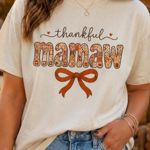 May include: A cream-colored short-sleeve t-shirt featuring the text thankful mamaw in a decorative font. The word mamaw is filled with a multi-colored floral quilt pattern. Below the text is a large, solid burnt orange bow graphic.