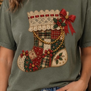 May include: An olive green t-shirt featuring a large graphic of a Christmas stocking. The stocking has a patchwork design in red, green, and cream, decorated with gold beads, a red ribbon bow, lace trim, and small holly berry accents.