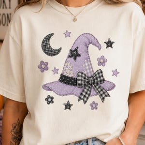 May include: A cream-colored short-sleeve t-shirt featuring a graphic of a purple witch hat with a black and white checkered bow. The design includes a crescent moon, various stars, and small flowers in shades of purple, black, and white.