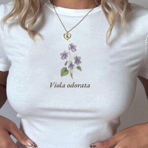 May include: White t-shirt featuring a watercolor illustration of purple violets and green leaves, with the text "Viola odorata" in brown. A gold heart-shaped pendant necklace hangs from the neckline.