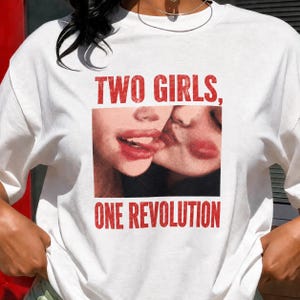 Può includere: T-shirt bianco con grafica di due labbra, una che lecca l'altra. Il testo rosso "TWO GIRLS, ONE REVOLUTION" è stampato sopra e sotto l'immagine. La maglietta è in stile casual.