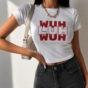 Puede incluir: Top corto blanco con el texto rojo "WUH LUH WUH". La camiseta tiene mangas cortas y cuello redondo. La modelo lleva vaqueros negros y un collar de cadena dorada.