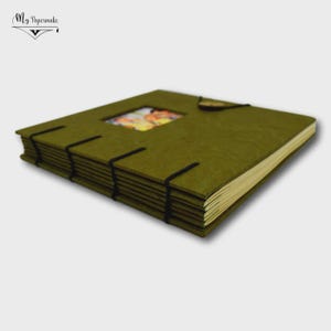 May include: A square photo album with a textured olive green cover featuring a central square cutout displaying a colourful image. The album is bound with black elastic cords that wrap around the spine and cover, holding the pages securely together.