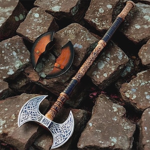 Può includere: Un'ascia a doppia lama con intricati disegni a nodo celtico sulle lame argentate e sul manico in legno. L'impugnatura è avvolta in pelle blu scuro. È presente anche un fodero in pelle, appoggiato su un letto di rocce.