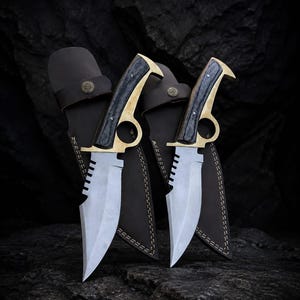 May include: Two fixed-blade knives with silver-toned steel blades featuring serrated spines, brass guards with integrated finger rings, and dark wood handles. Each knife is paired with a dark brown leather sheath featuring a brass button stud.