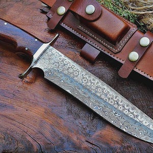 May include: A Damascus steel knife with a dark wood handle and a brown leather sheath. The blade has a unique patterned design. The sheath has a belt loop and snap closure. The knife is on a wooden surface.