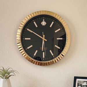 May include: A round wall clock with a gold fluted frame and a black face featuring gold hour markers, gold hands, and a small gold crown icon at the twelve o'clock position. The clock displays the text "QUARTZ" and "LONDON" in small gold lettering.