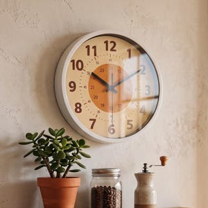 Peut inclure: Une horloge murale ronde avec un cadre blanc, un cadran crème et un cercle central orange avec des chiffres et des aiguilles noirs. En dessous, une étagère en bois porte une plante verte dans un pot en terre cuite, un bocal en verre et un moulin.