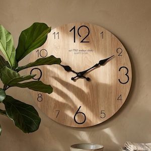 May include: A round wall clock with a light wood grain finish, black numerals, and black metal hands. The clock face features the text "est 1765 vintage clocks QUARTZ CLOCK" printed in black near the centre, positioned below the number 12.