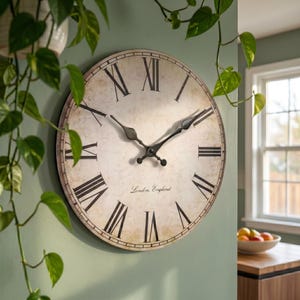 May include: A round wall clock with a distressed cream-coloured face, black Roman numerals, and black metal hands. The centre of the clock features the text London, England in a script font. The clock has a thin dark frame and a weathered finish.