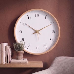 Op de afbeelding: Een ronde wandklok met een lichthouten frame, een witte wijzerplaat en zwarte Arabische cijfers van 1 tot 12. De klok heeft dunne goudkleurige wijzers voor uren, minuten en seconden, met kleine zwarte streepjes voor de minuten.