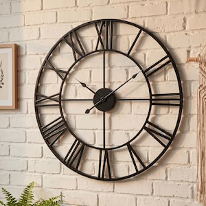 May include: A large, round, black metal wall clock with Roman numerals. The clock has a minimalist design with a circular frame and black hands. The clock is mounted on a white brick wall, adding a touch of industrial style to the decor.
